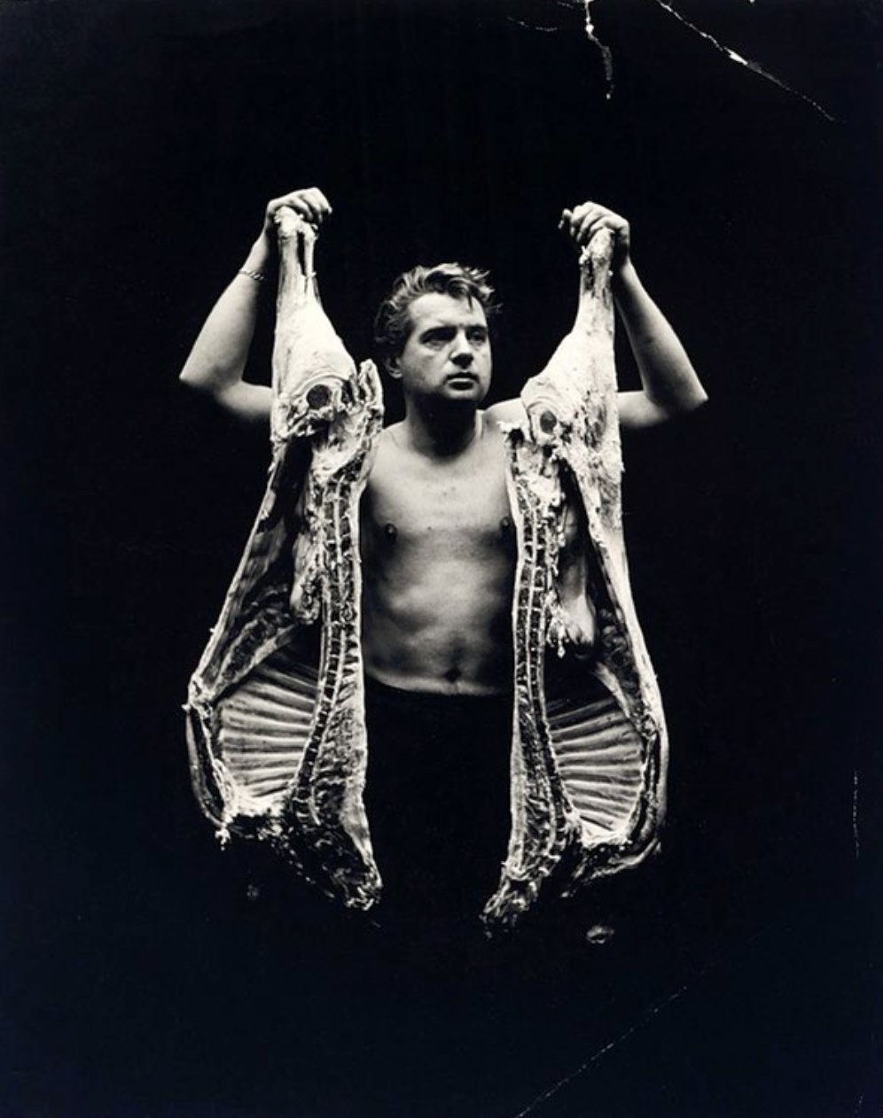 Francis Bacon torse nu, tenant à bras levés deux demi-carcasses de bœuf, photographie noir et blanc