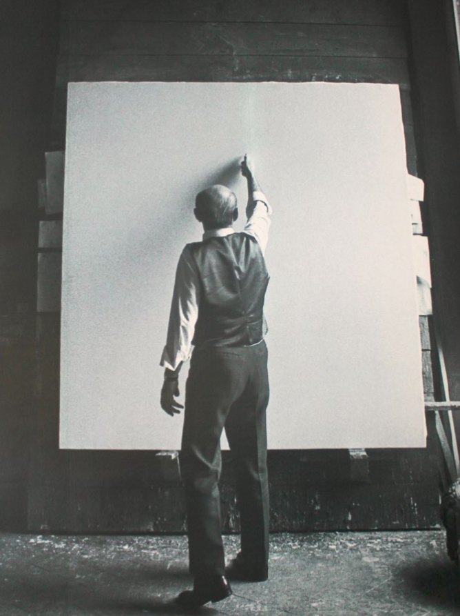 Lucio Fontana de dos, bras levé, devant une grande toile blanche